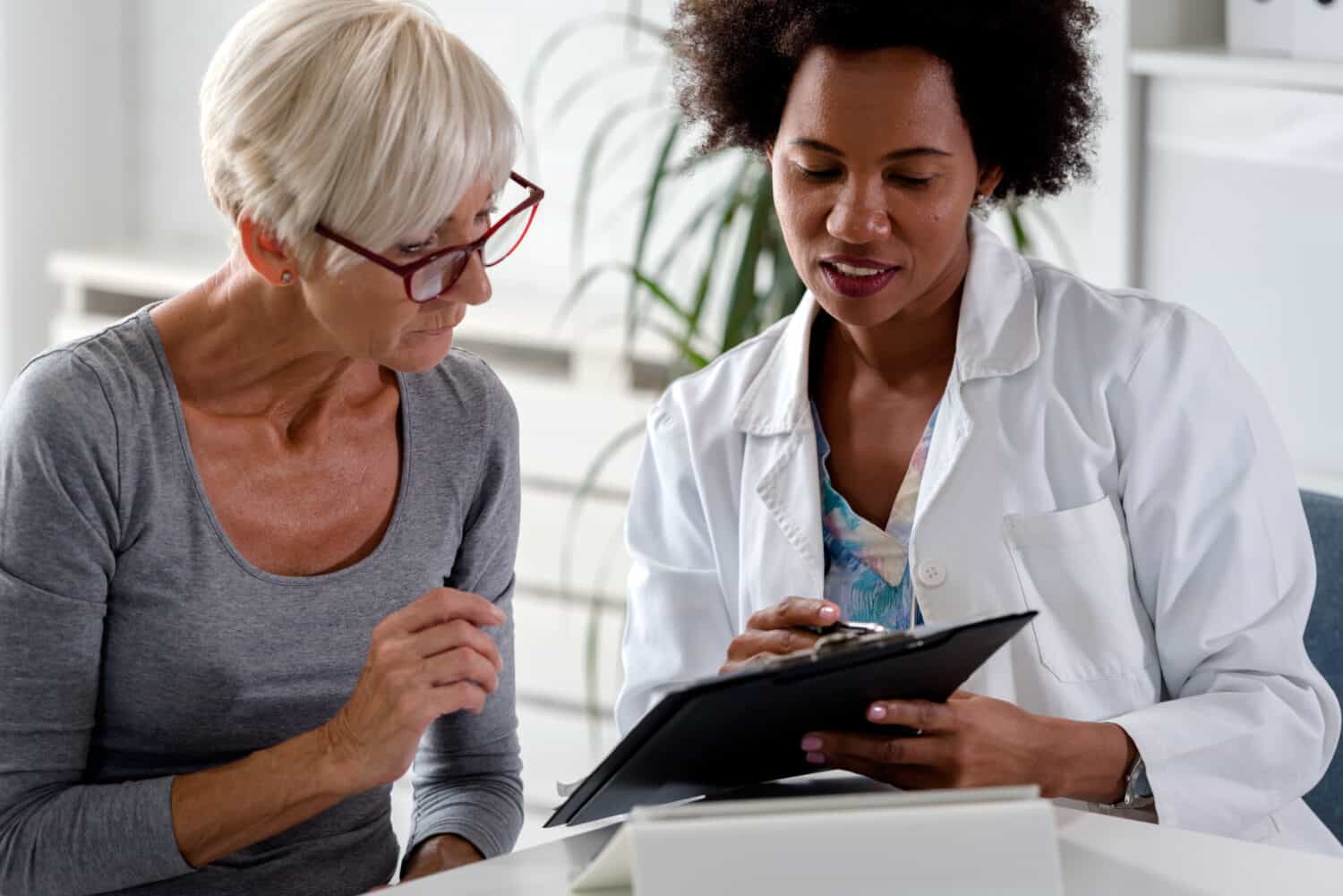 A female health practitioner and hearing aid expert sits at a table with a patient to discuss hearing loss, the best free online hearing test and a hearing aid price.