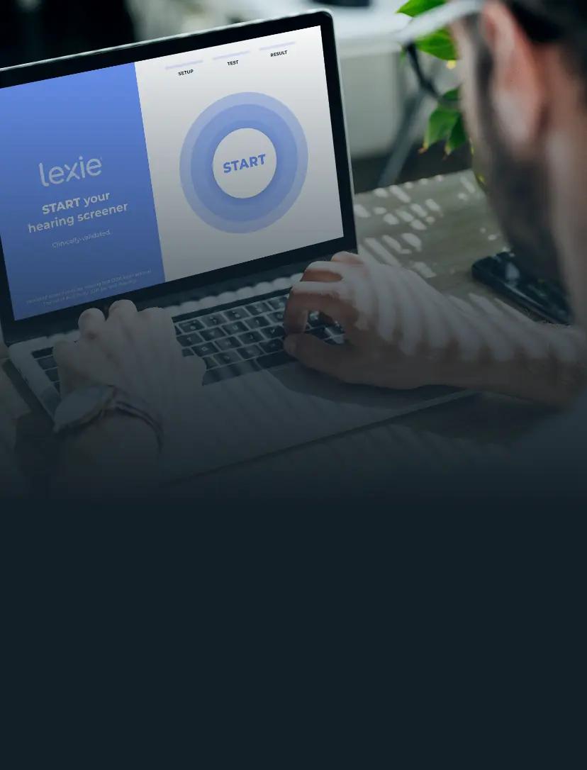 Man wearing glasses sitting at a table with his laptop open. On the laptop screen is Lexie Hearing’s free online hearing test.