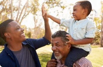 A father, a grandfather and a son outside in a park. All 3 people are smiling.